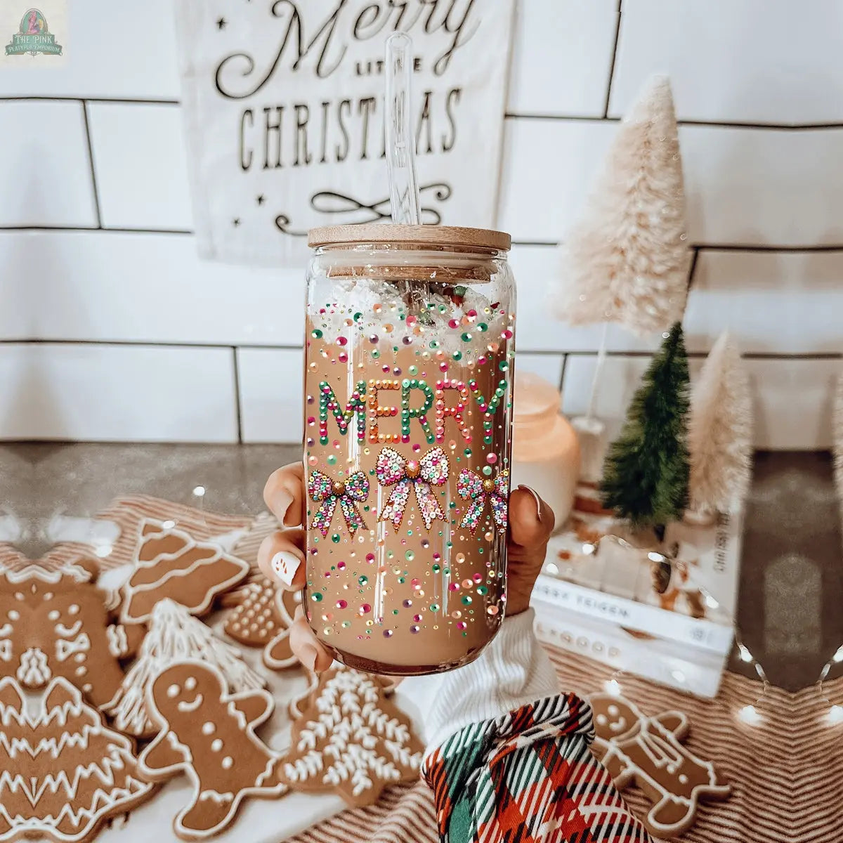 A hand holds a Sequin MERRY holiday glass tumbler of iced coffee topped with whipped cream and sprinkles. Gingerbread cookies, Christmas decor, and miniature trees decorate the festive background.