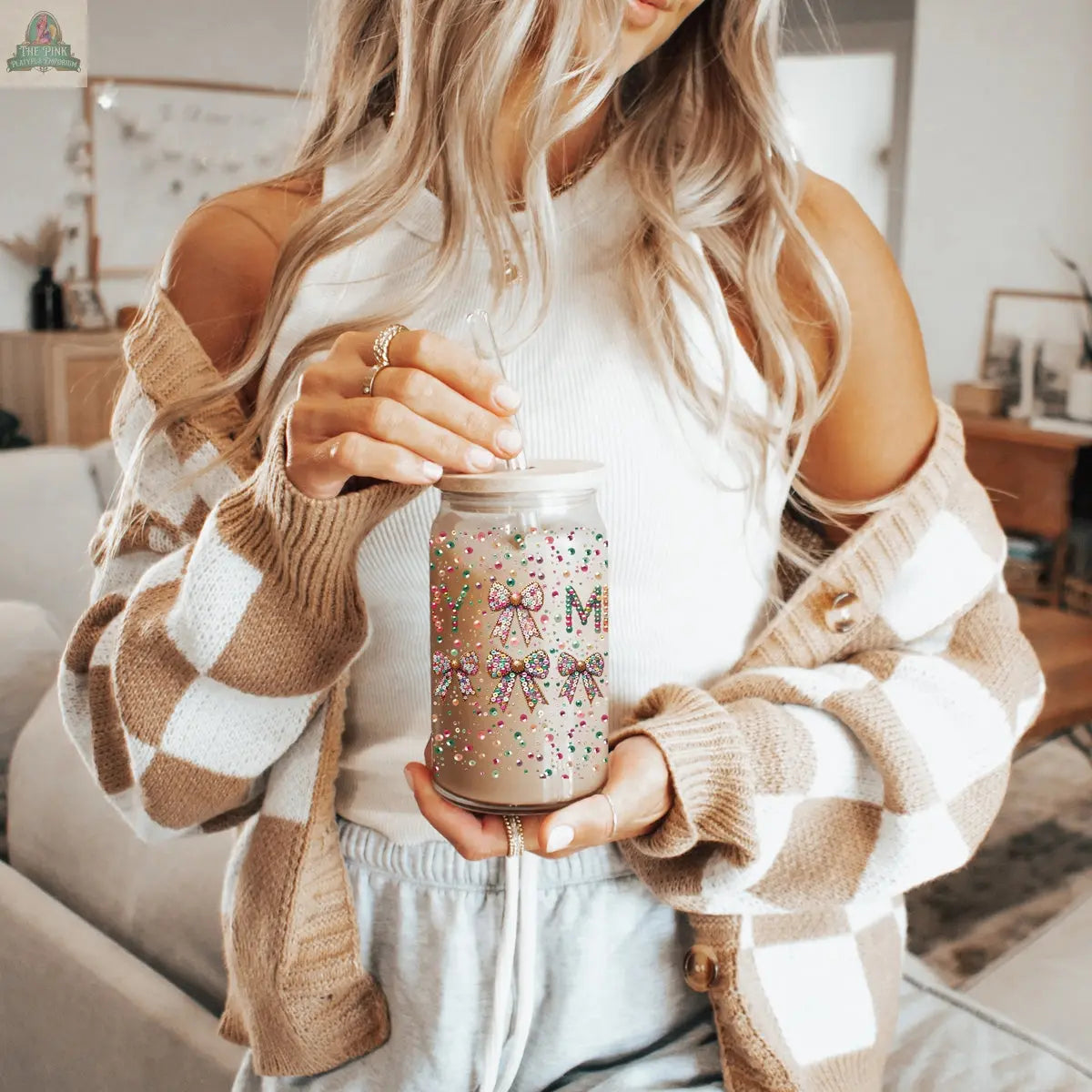 A woman with long wavy blonde hair holds the Sequin MERRY holiday glass tumbler with a lid and straw, decorated with colorful sprinkles. She wears a white sleeveless top, brown plaid cardigan, and light gray shorts.