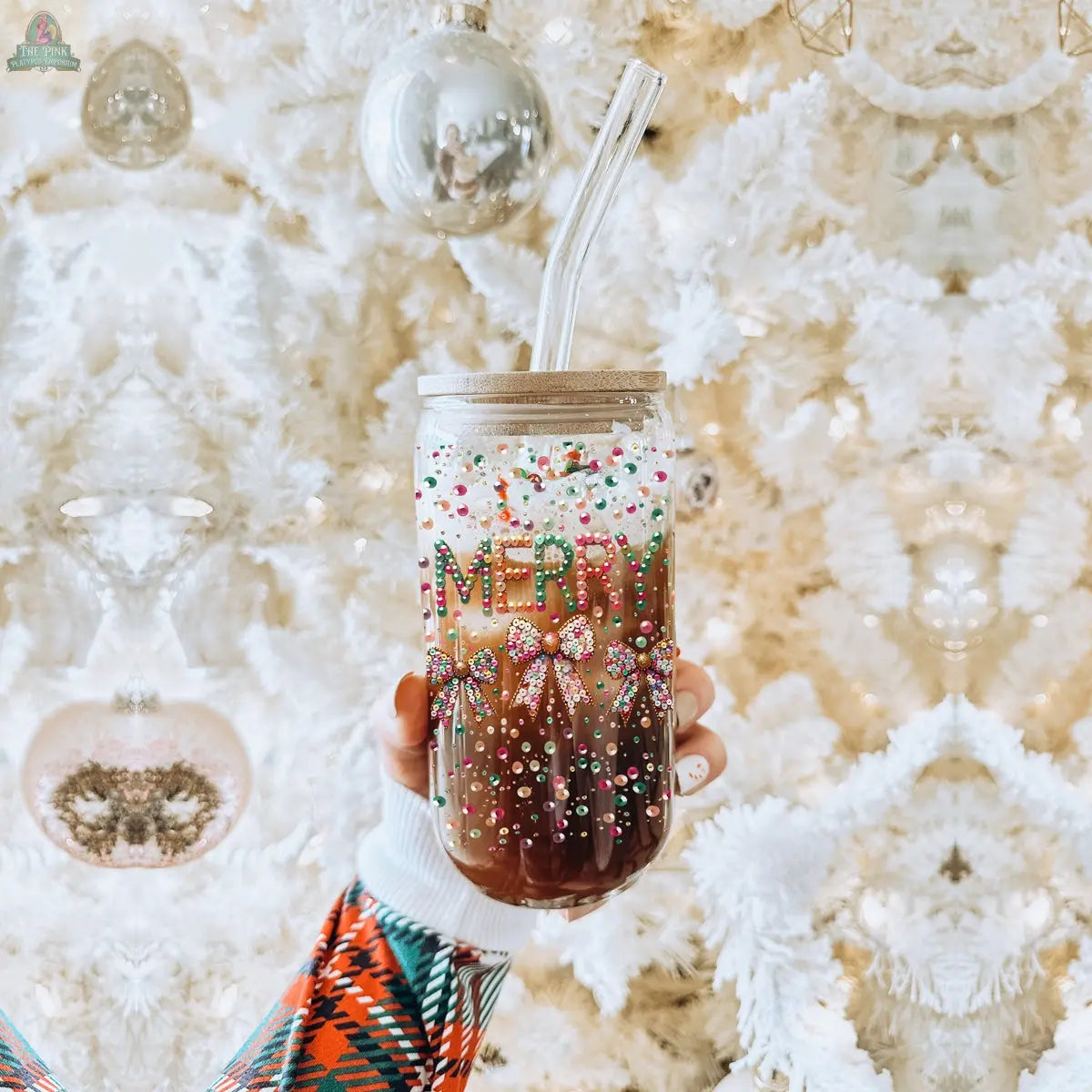 A hand holds the Sequin MERRY tumbler glass cup with colorful beads, sprinkles, a clear straw and lid. In the background, a white Christmas tree with ornaments enhances the festive charm of this holiday tumbler glass.