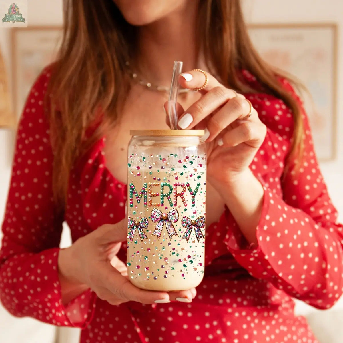 A woman in a red polka dot dress holds the Sequin MERRY glass tumbler, filled with a creamy drink topped with rainbow sprinkles, sipping through a straw.