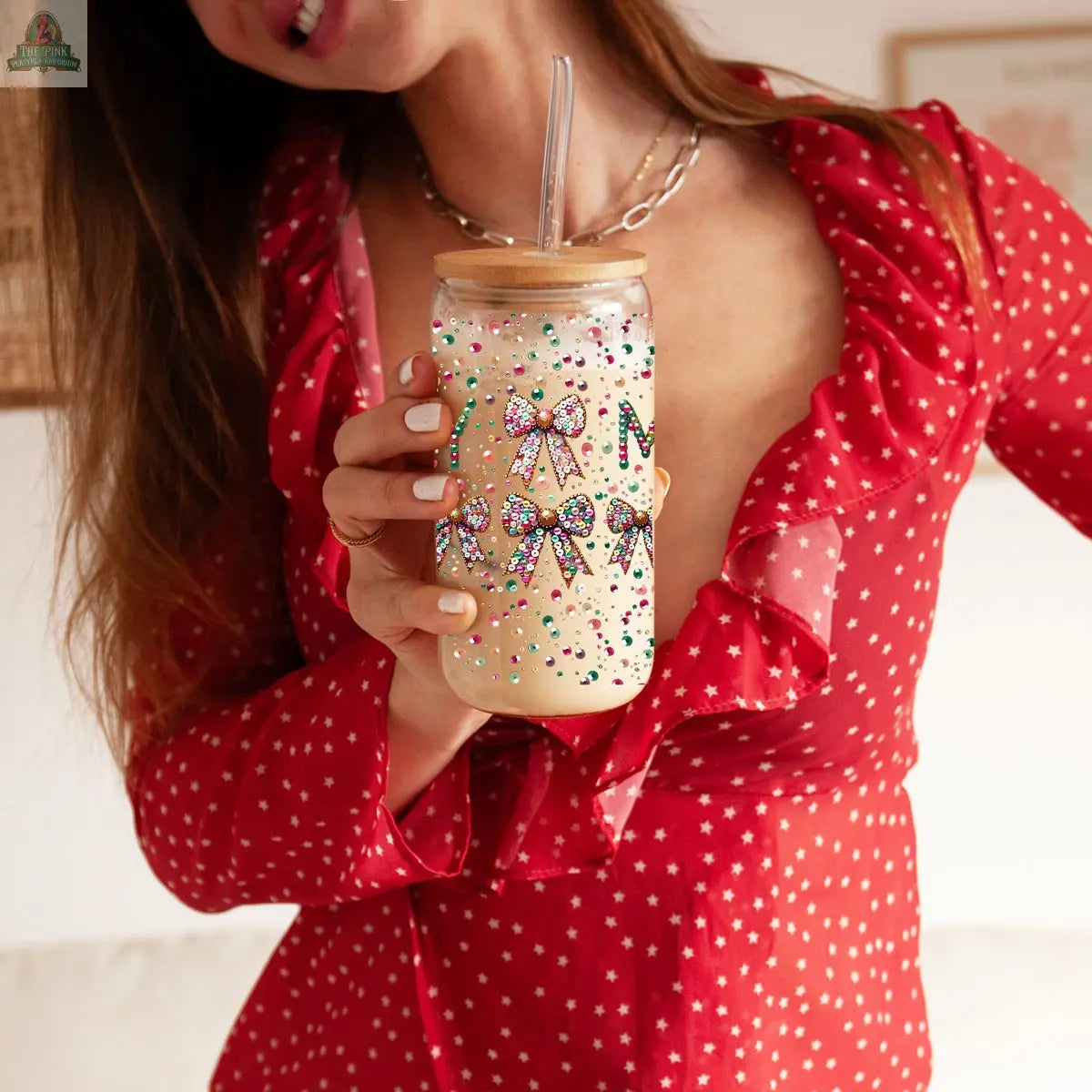 A woman in a red, star-patterned dress holds the Sequin MERRY holiday mason jar glass tumbler, decorated with colorful beads, bows, and a straw. It contains a creamy drink; her face is partially out of frame.