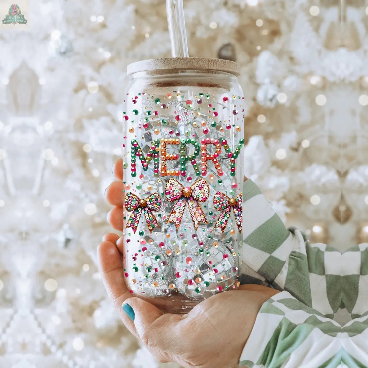 A hand holds the Sequin MERRY tumbler, a holiday glass covered in colorful rhinestones spelling "MERRY" and three sequin bows. A clear straw extends from the top, with a softly glowing white Christmas tree in the background.