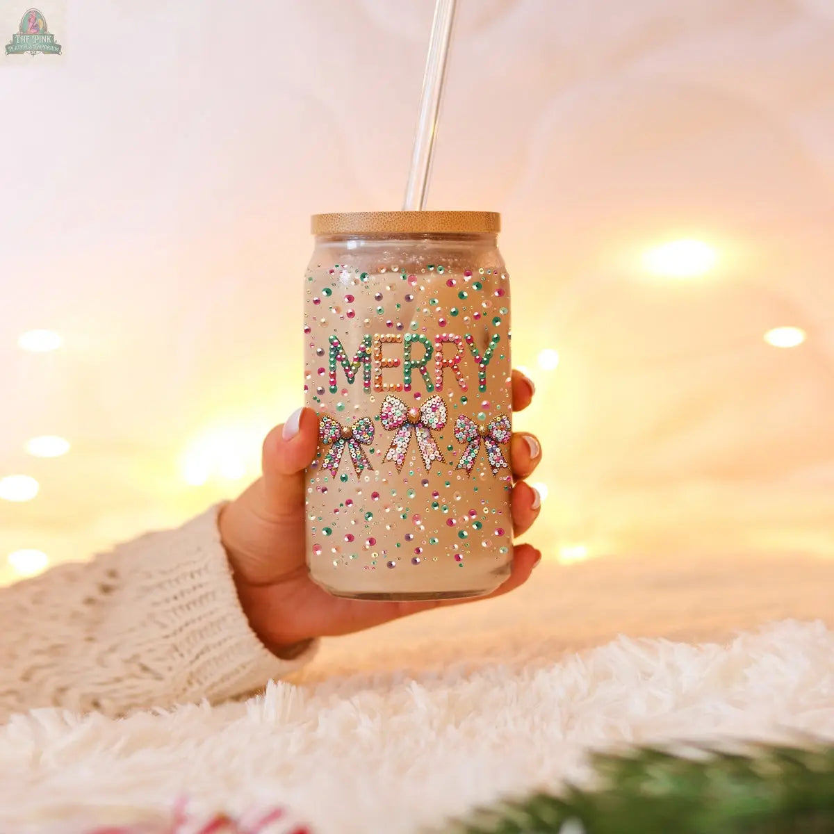 A hand holds the Sequin MERRY glass tumbler with a straw, filled with a light brown drink. The tumbler features colorful beads spelling MERRY and sequin bows, with warm fairy lights softly glowing in the background.