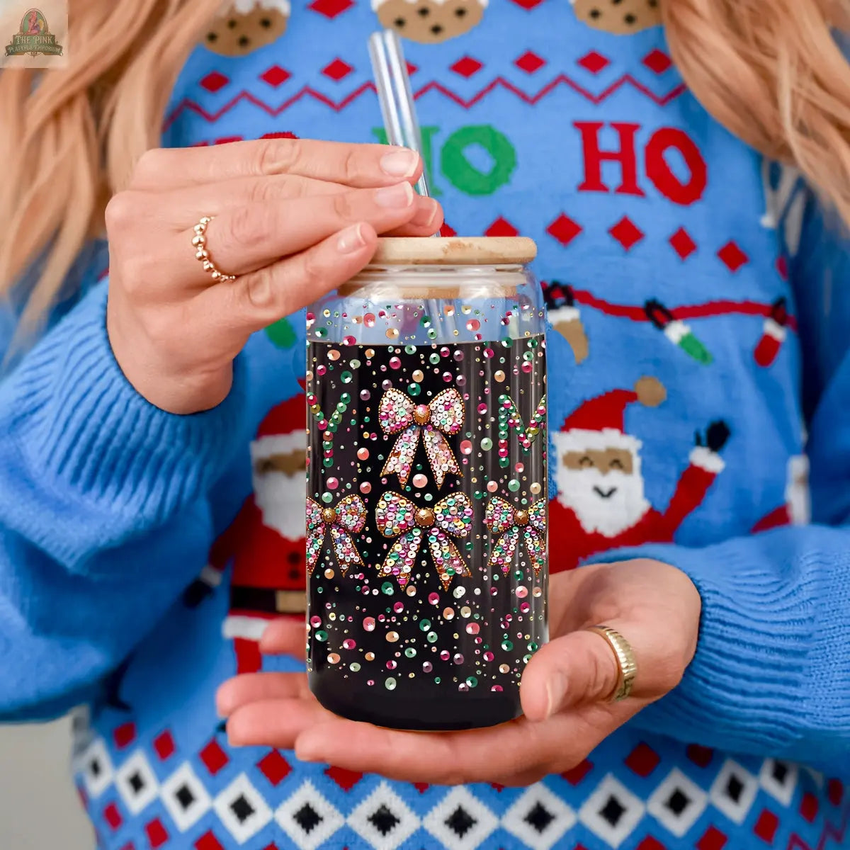 Wearing a blue holiday sweater with "HO HO HO" and Santa Claus designs, a person holds the Sequin MERRY tumbler—a glass jar decorated with colorful rhinestone bows, pearls, and finished with a wooden lid and straw.