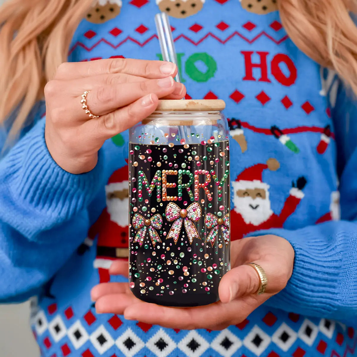 Wearing a festive blue Christmas sweater, a person holds the Sequin MERRY tumbler—a dazzling holiday glass adorned with sequined gems spelling "MERRY" and bows. Their painted nails and sparkling ring add extra festive flair.