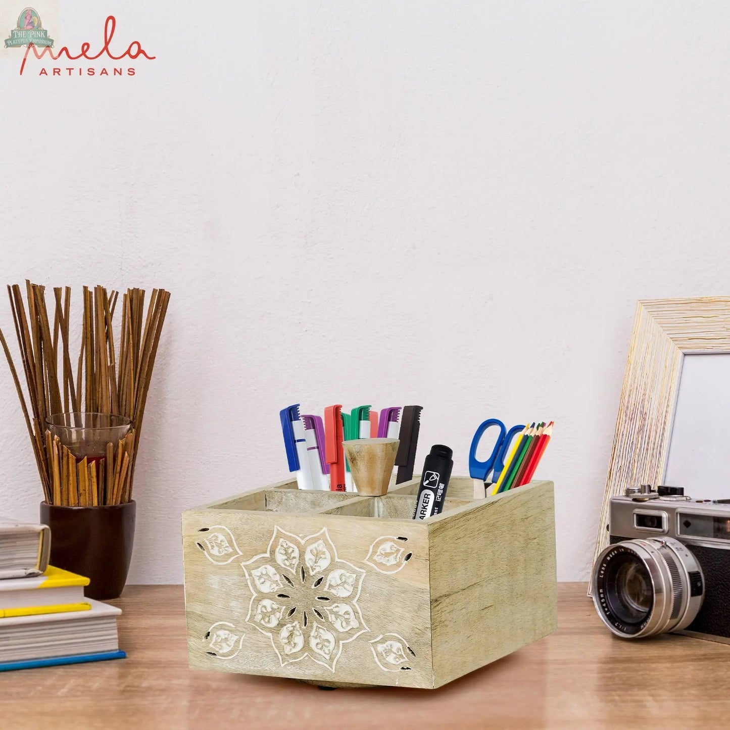 The Serena Spinner 4 Quad in Whitewash keeps your pens and markers organized on your desk, next to a camera, photo frame, books, and a bundle of sticks. "mela ARTISANS" is displayed boldly in red on the wall behind.
