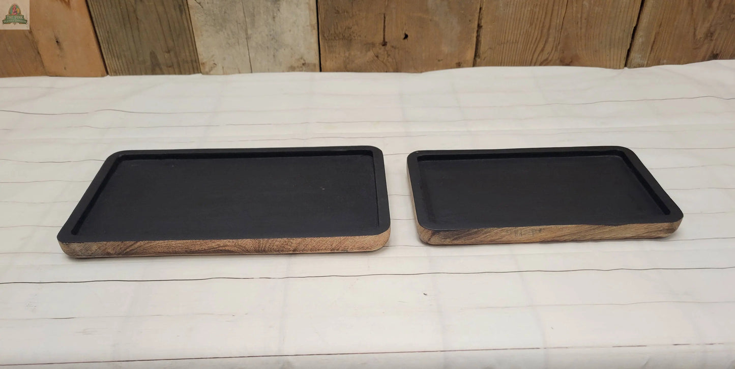 Two Wood Trays with a matte black finish and wooden sides rest on a white striped cloth. The larger tray is on the left of the smaller, both placed on rustic wooden planks.
