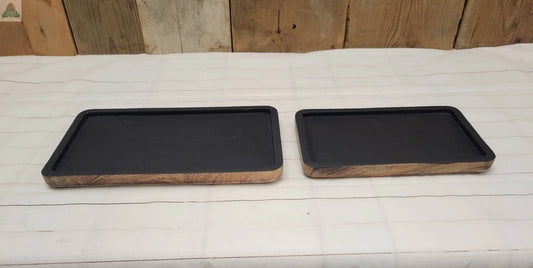 Two Wood Trays with a matte black finish and wooden sides rest on a white striped cloth. The larger tray is on the left of the smaller, both placed on rustic wooden planks.