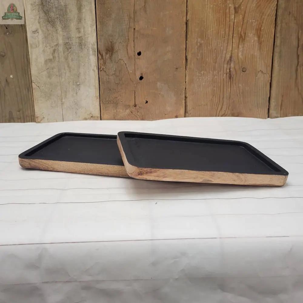 Two Wood Trays with a matte black finish are displayed on white fabric against a rustic wood wall—one tray lies flat while the other stands angled, adding a modern yet timeless accent to your decor.