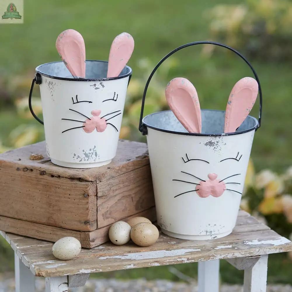 The Set of Two Bunny Buckets features white metal buckets with painted bunny faces and pink ears, displayed outdoors on wood with three small eggs nearby—ideal as charming Easter planters amid lush green foliage.