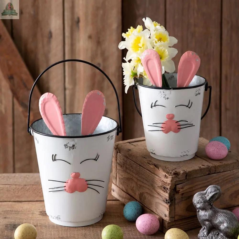 The Set of Two Bunny Buckets features charming white planters with pink ears, holding yellow flowers on wooden surfaces. Colorful Easter eggs and a silver bunny accent complete these rustic Easter decorations.