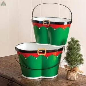 A Set of Two Elf Pants Buckets—green metal nesting containers with black handles, red trim, and gold buckle accents—are stacked on a wooden surface beside a small potted artificial pine tree.