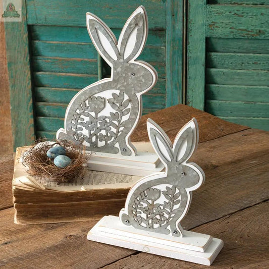The Set of Two Wooden Bunnies with Metal Cutouts sits near a bird's nest with blue eggs, displayed on a wooden surface against green shutter panels and an old book for a charming rustic look.