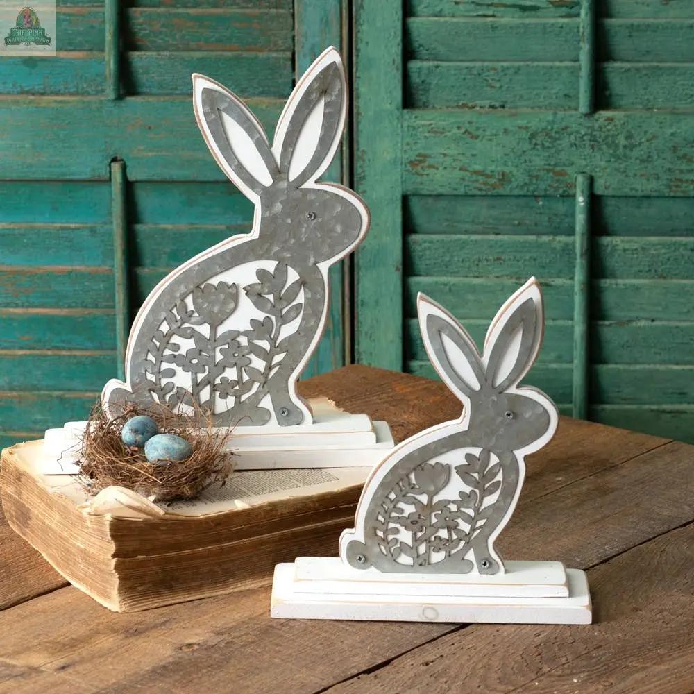 A Set of Two Wooden Bunnies with Metal Cutouts sits on white bases atop a wooden table, accompanied by a bird’s nest with blue eggs and green wooden shutters in the background.