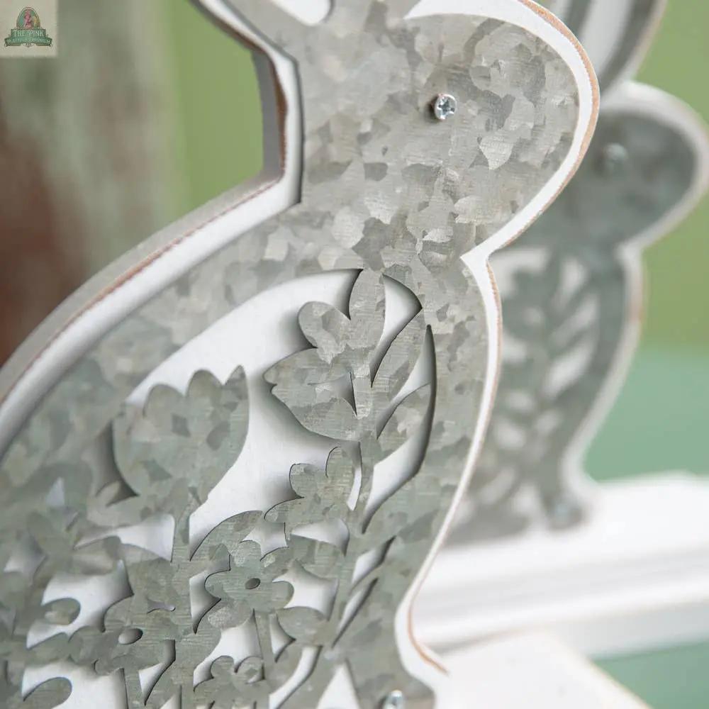 Close-up of a bunny from the Set of Two Wooden Bunnies with Metal Cutouts, featuring a decorative floral metal design on a white wooden base. The second bunny from the set appears slightly in the background.