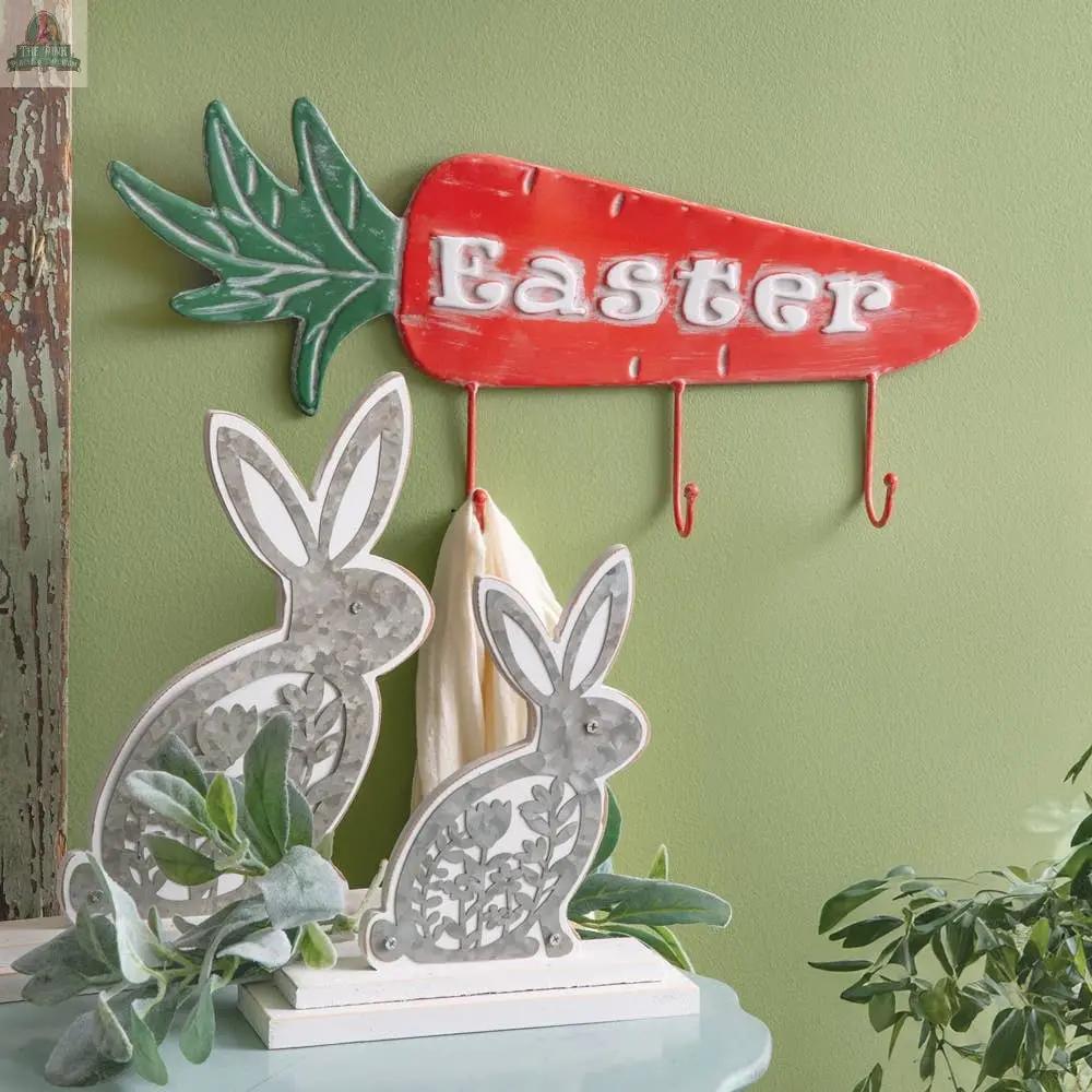 Decorative Easter display featuring a large carrot-shaped "Easter" sign with three hooks and a Set of Two Wooden Bunnies with floral metal cutout details, accented by green foliage against a light green wall.