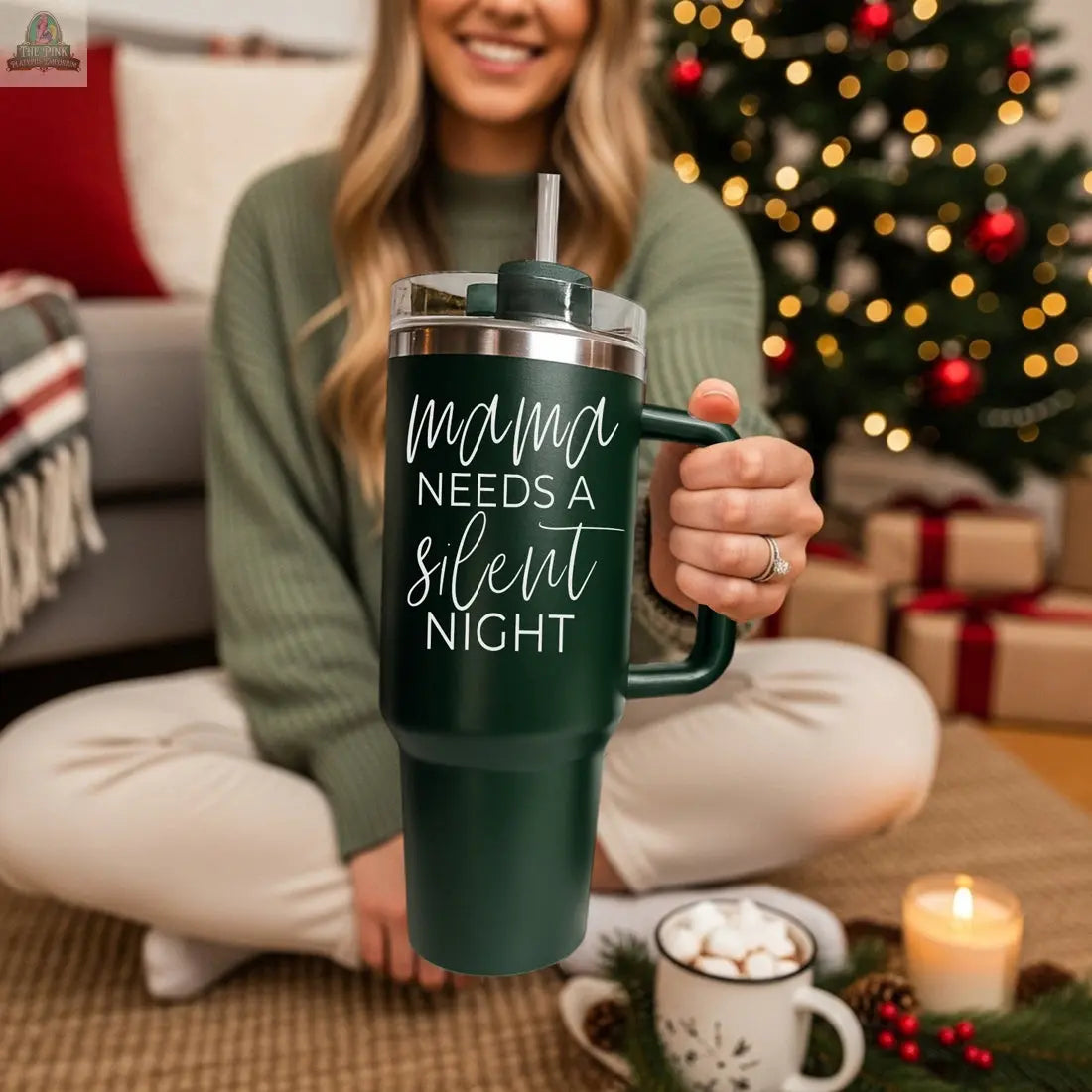 A woman sits cross-legged by a decorated Christmas tree, holding the Silent Night 40oz tumbler. Wrapped gifts, a lit candle, and a mug of hot chocolate with marshmallows are beside her.