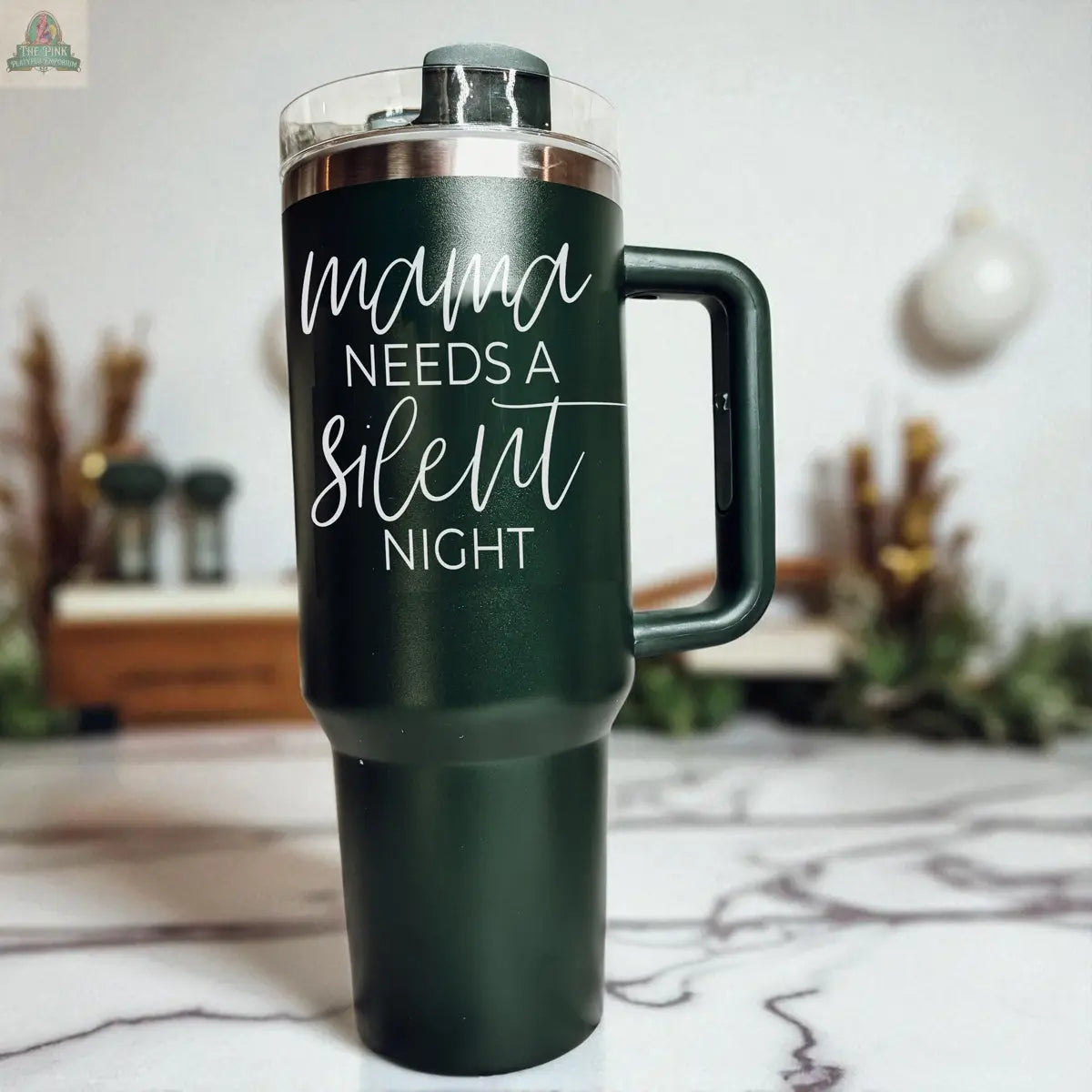 A dark green Silent Night 40oz insulated mug with a handle sits on a marble counter, printed with "mama needs a silent night" in white script. A blurred kitchen with holiday decor is in the background, making it a perfect Christmas gift.
