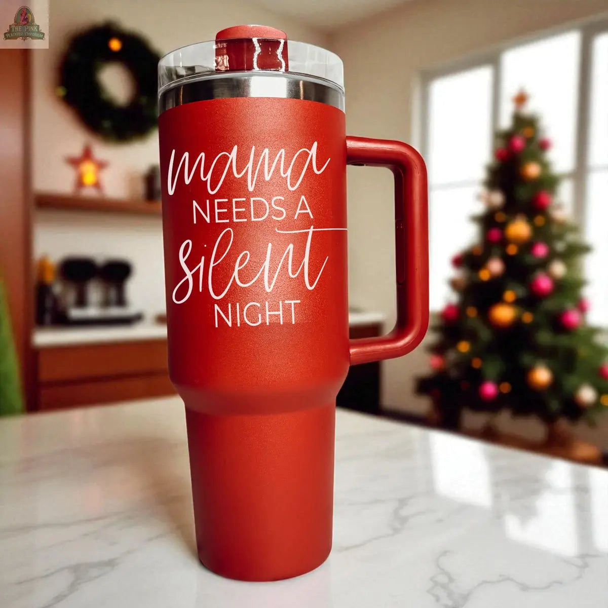 The Silent Night 40oz tumbler, featuring "Mama needs a silent night" in red, rests on a kitchen counter with a Christmas tree and holiday wreath in the background—perfect as a festive Christmas gift.