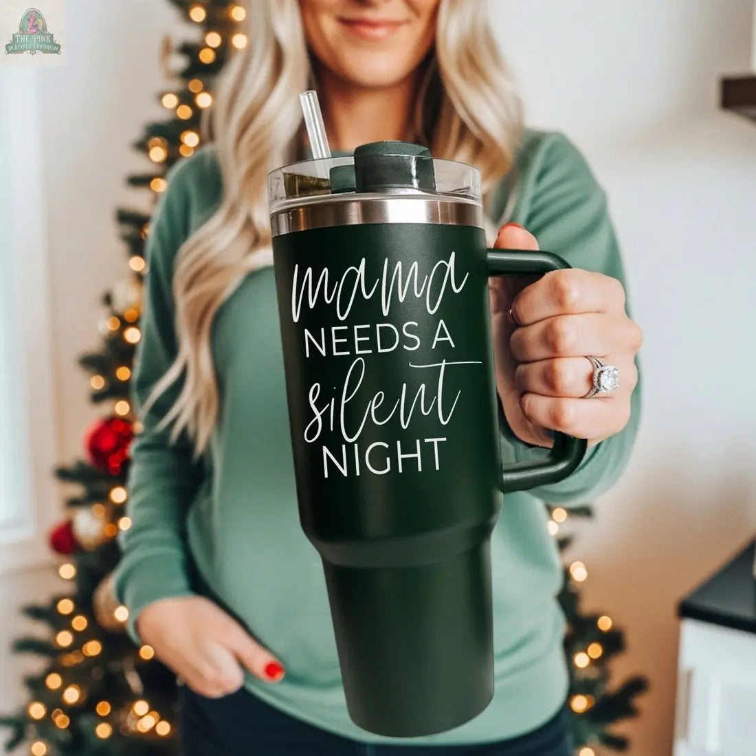 A woman in a green top stands by a decorated Christmas tree, holding the Silent Night 40oz tumbler featuring "Mama needs a silent night" in white script—a perfect insulated mug gift for the holidays.
