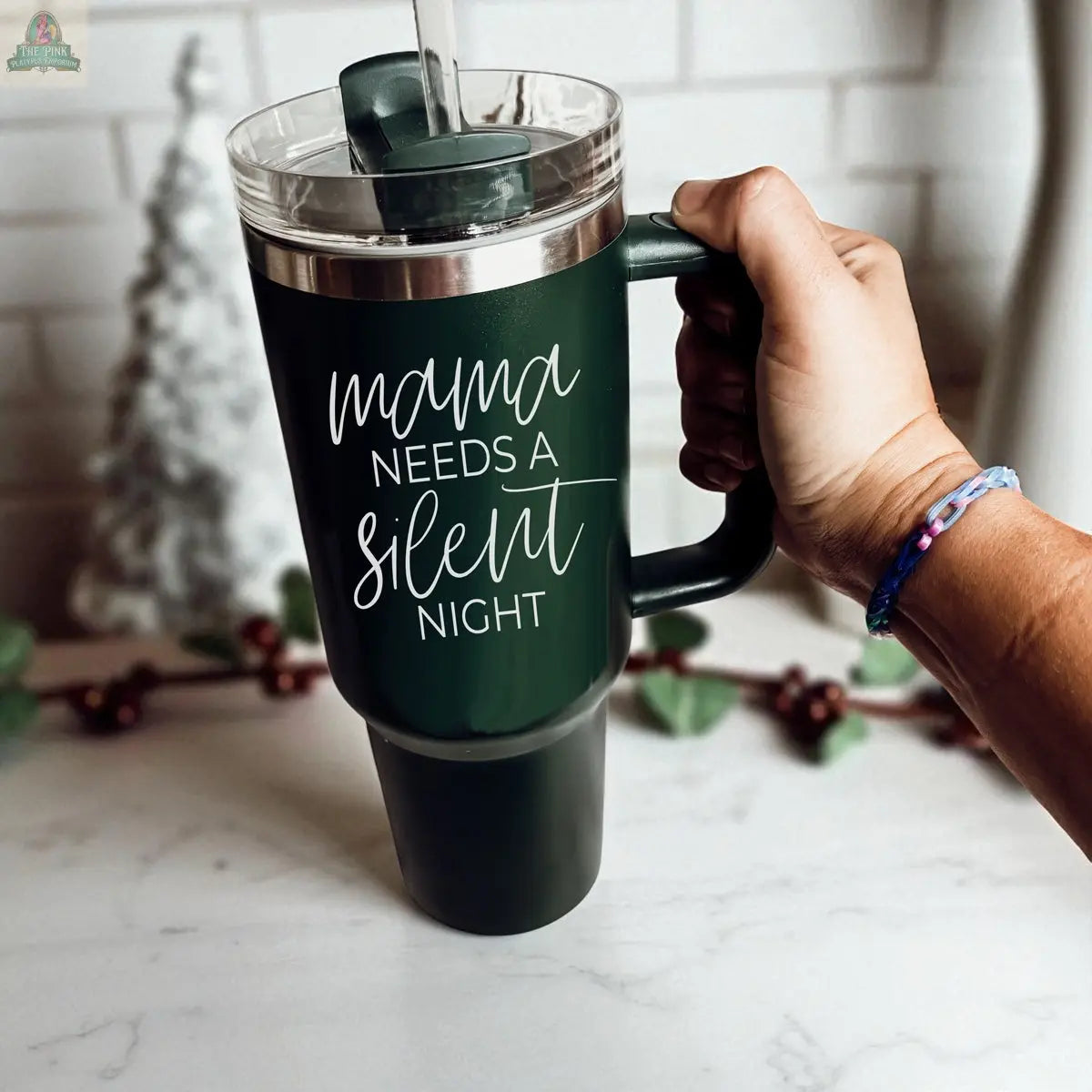 A hand holds the Silent Night 40oz tumbler with a straw, featuring "Mama needs a silent night" in white script. Its dark green color and festive design make it an ideal Christmas gift, with a blurred tree and garland in the background.
