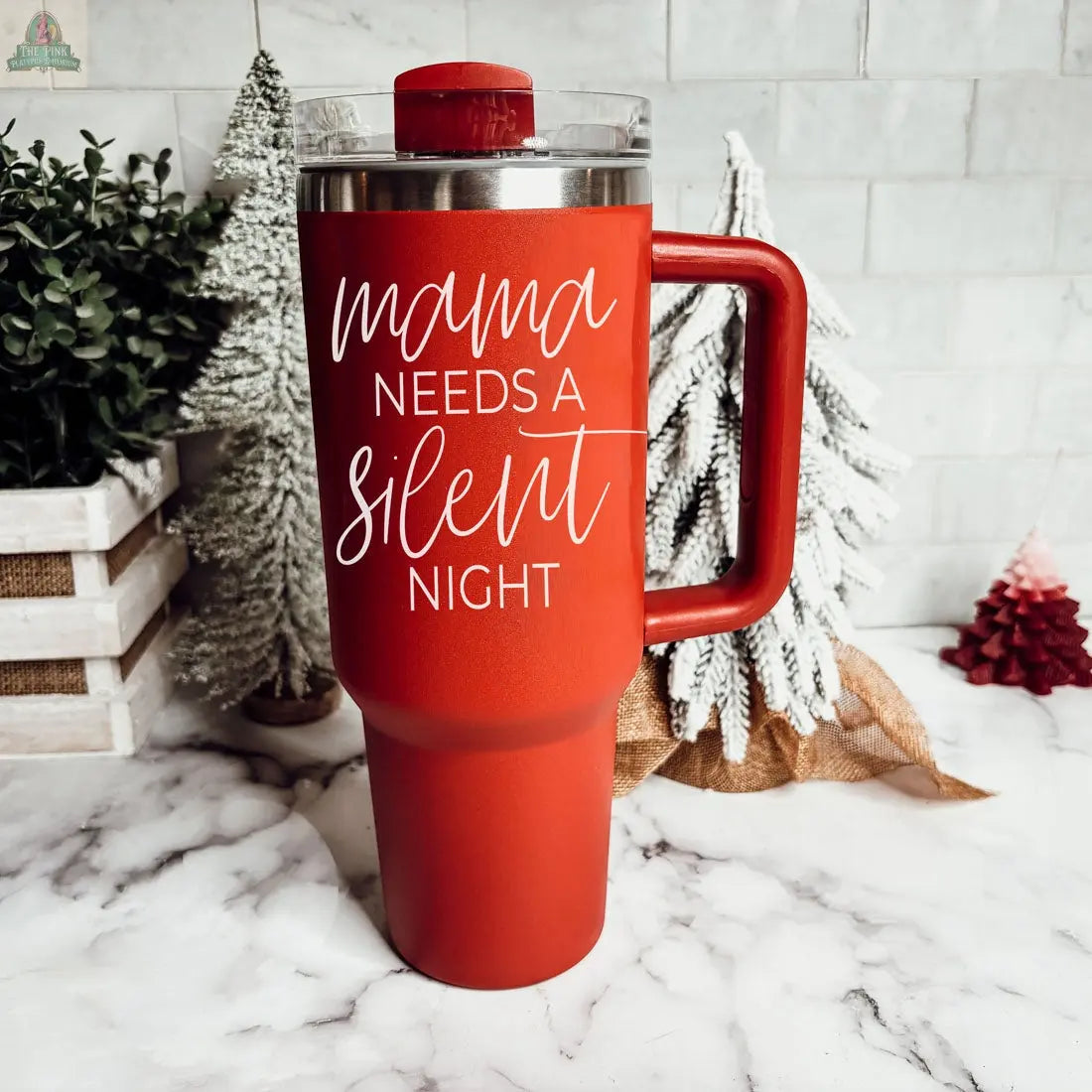 The Silent Night 40oz, a tall red insulated mug with a large handle and clear lid, sits on a marble counter reading “mama needs a silent night.” Snowy decorative trees and greenery in the background make it a perfect Christmas gift.