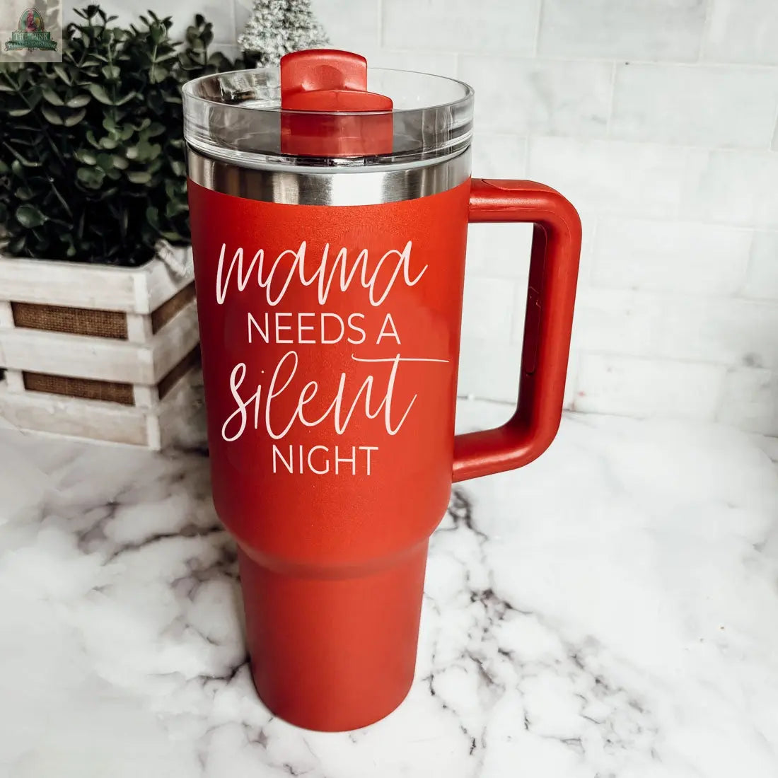The Silent Night 40oz tumbler, in red with a handle and clear lid, features “Mama needs a silent night” in white text—making it an ideal Christmas gift. It sits on a marble counter with plants and a wooden box in the background.