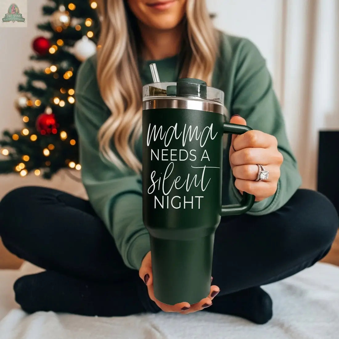 A woman with long blonde hair sits cross-legged by a decorated Christmas tree, holding the Silent Night 40oz tumbler in dark green, featuring the phrase "mama needs a silent night" in white script.