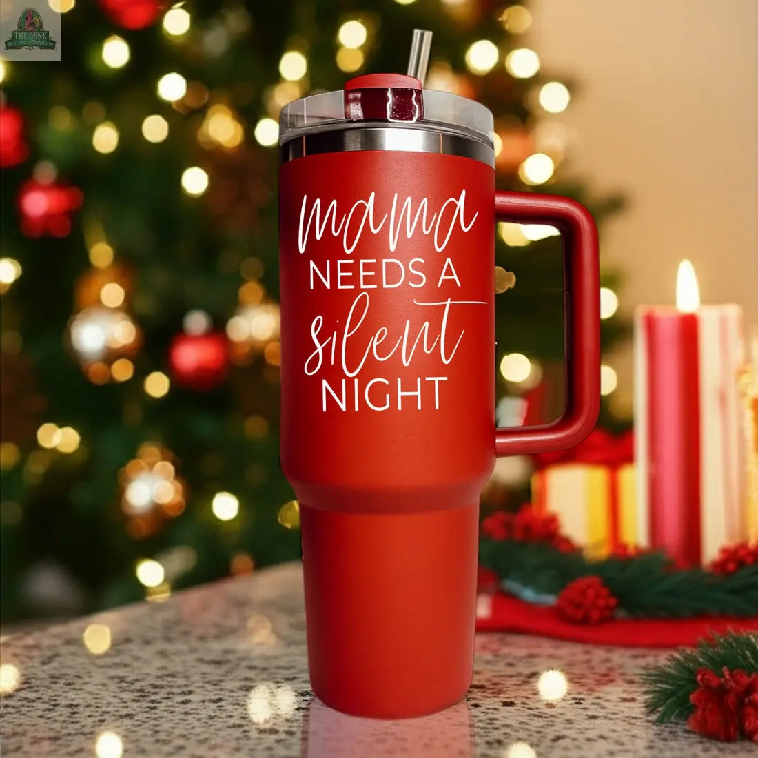 A red Silent Night 40oz insulated mug with a straw sits on a festive table, featuring "mama needs a silent night" in white text. Blurred Christmas lights, candles, and a decorated tree make it a perfect holiday gift.