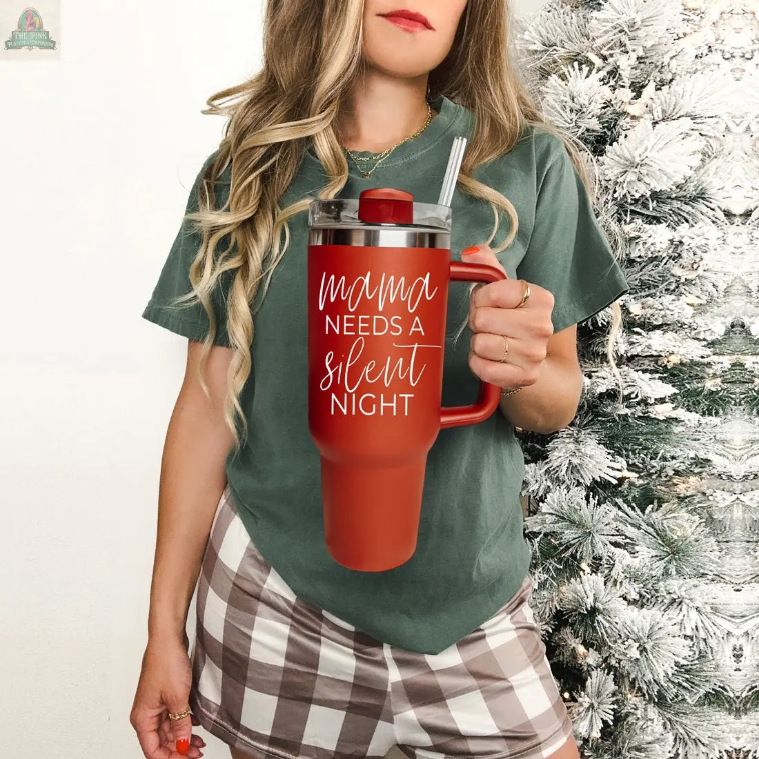 A woman in a green t-shirt and plaid shorts holds the Silent Night 40oz tumbler, featuring "Mama needs a silent night," beside a snowy Christmas tree—an ideal holiday gift for moms who love insulated mugs.