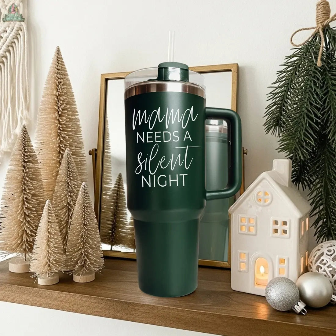 A Silent Night 40oz dark green tumbler sits on a wooden shelf, surrounded by faux pine trees, a lit ceramic house, and a pine branch—perfect for Christmas. A mirror is propped behind the tumbler.