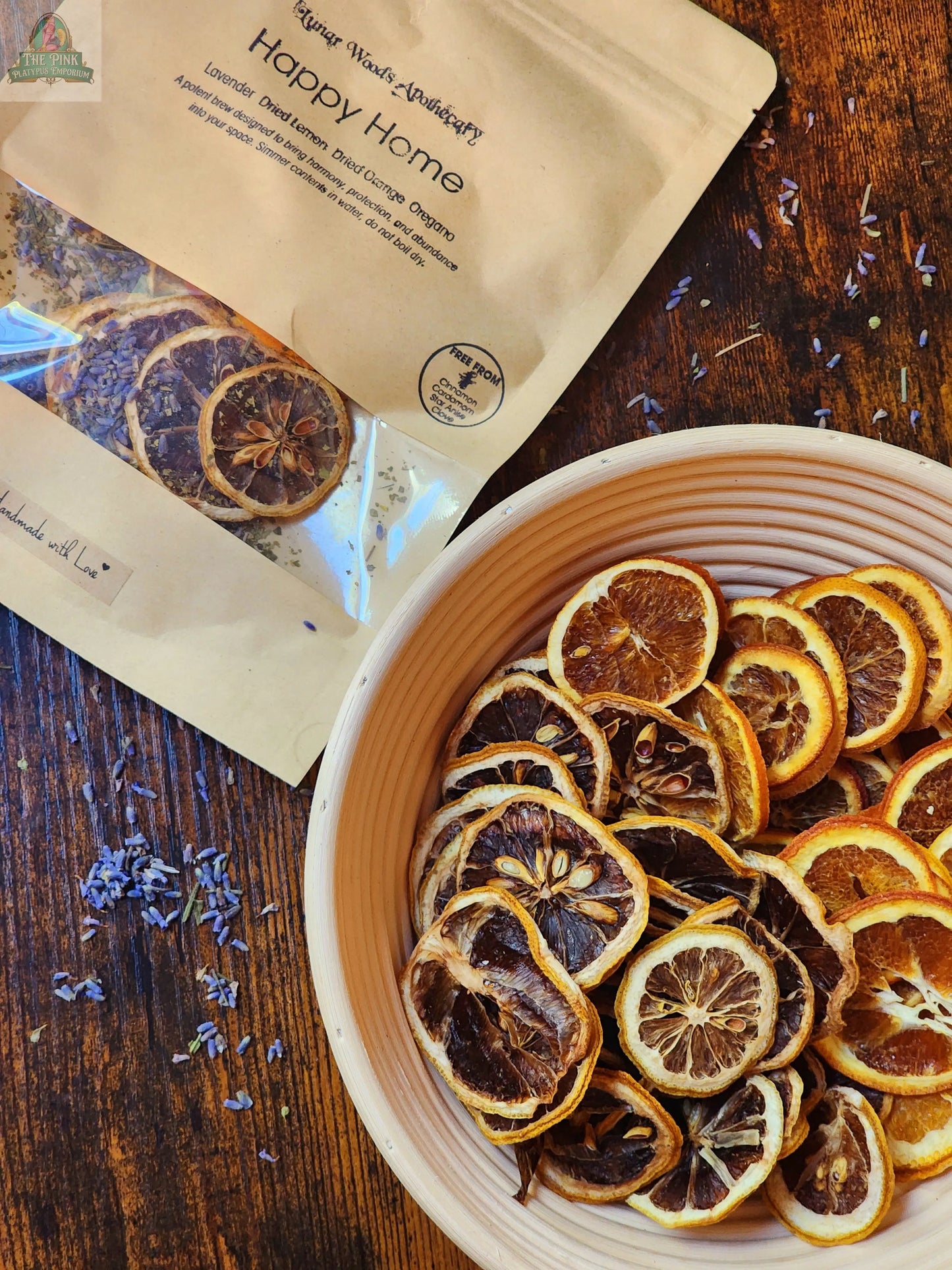 A bowl of dried orange slices and scattered lavender buds sits beside the Simmer Pot Kit- Happy Home beige packet on a wooden table, creating a charming and cozy home fragrance display.