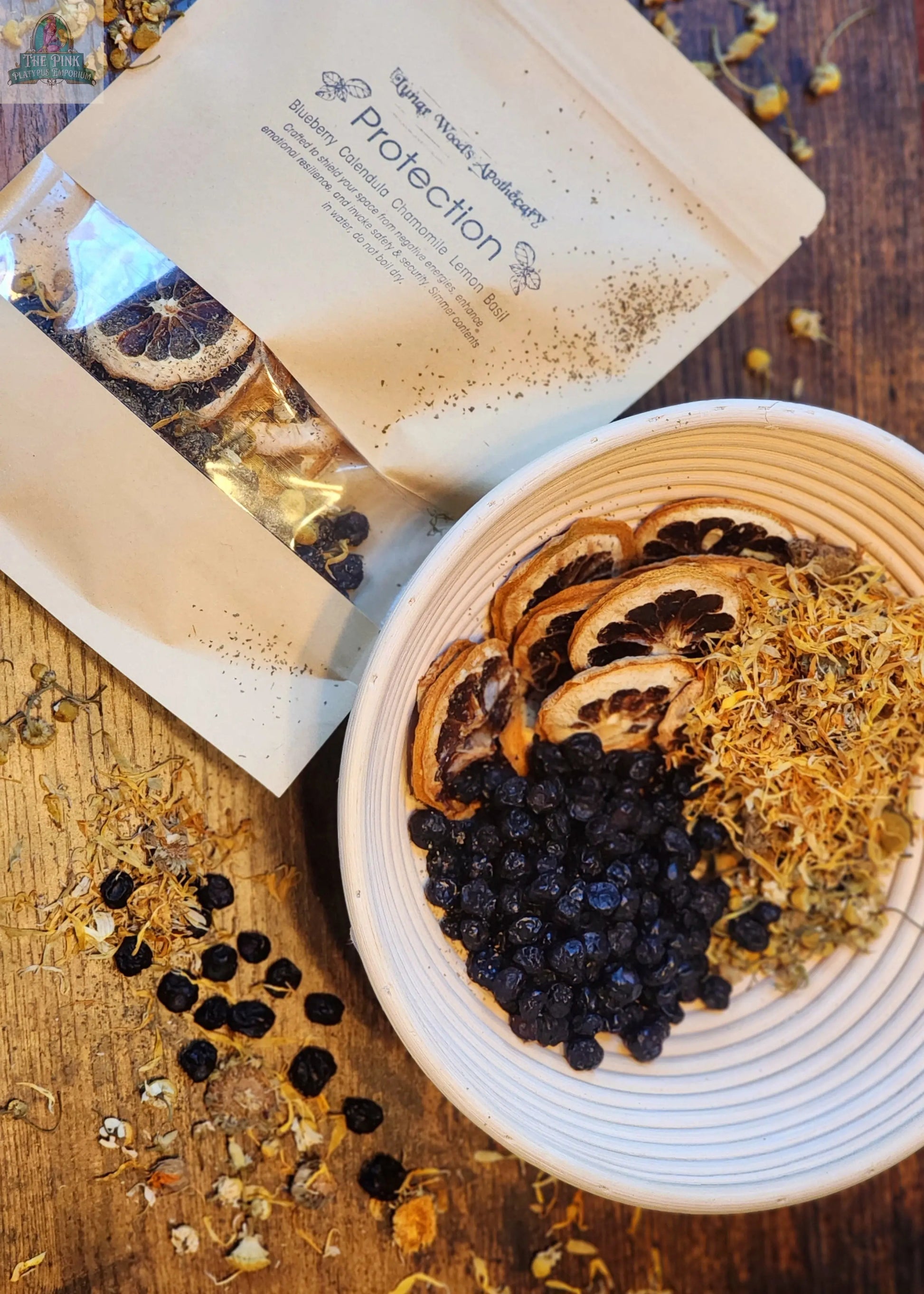 A bowl of dried citrus, blueberries, and herbs sits beside a Simmer Pot Kit- Protection packet with similar botanicals—ideal for cleansing your space. Loose berries and herbs are scattered on the wooden surface.