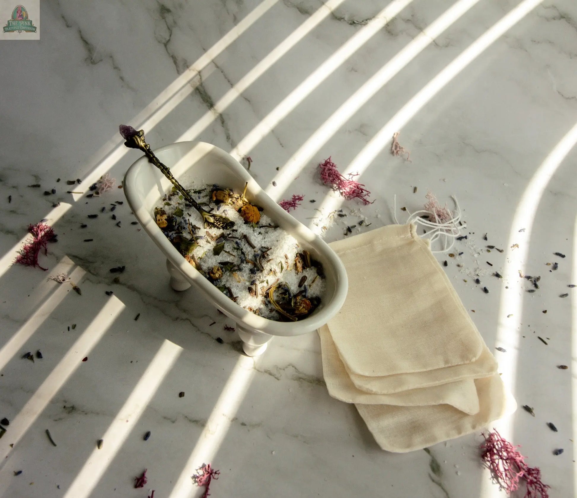 Sleep Well—a small bathtub-shaped dish with bath salts and dried flowers for a soothing herbal bath—rests on marble beside cream drawstring bags and scattered petals, inviting relaxation under striped sunlight.