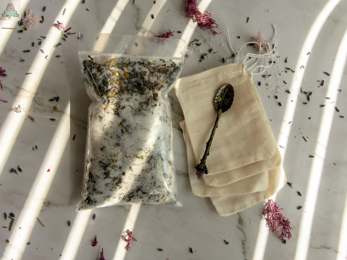 Sleep Well: Sealed bag of bath salts with dried herbs for restful sleep, vintage spoon, empty muslin bags, and scattered petals on a sunlit marble surface.