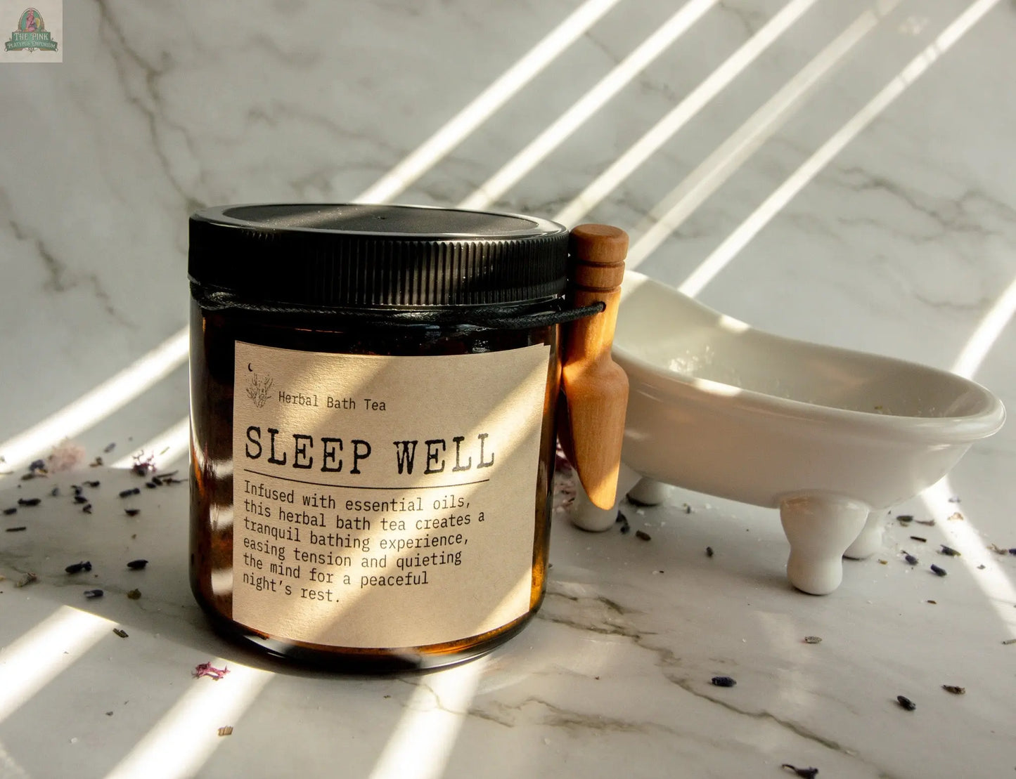 A jar of Sleep Well bath tea rests next to a white dish and wooden scoop, promising relaxation as sunlight filters through blinds, casting striped shadows on the marble surface.