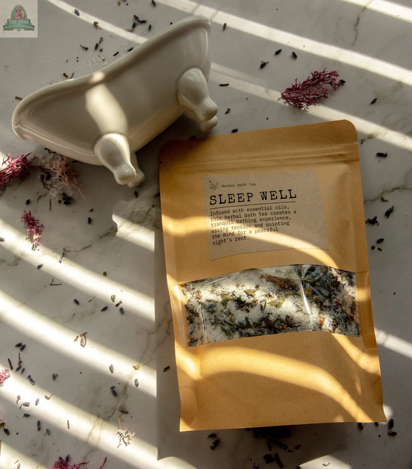 A packet of Sleep Well bath tea sits on a sunlit marble surface next to a white tub-shaped dish, surrounded by dried flowers and herbs, creating an inviting atmosphere for deep relaxation and restful sleep.