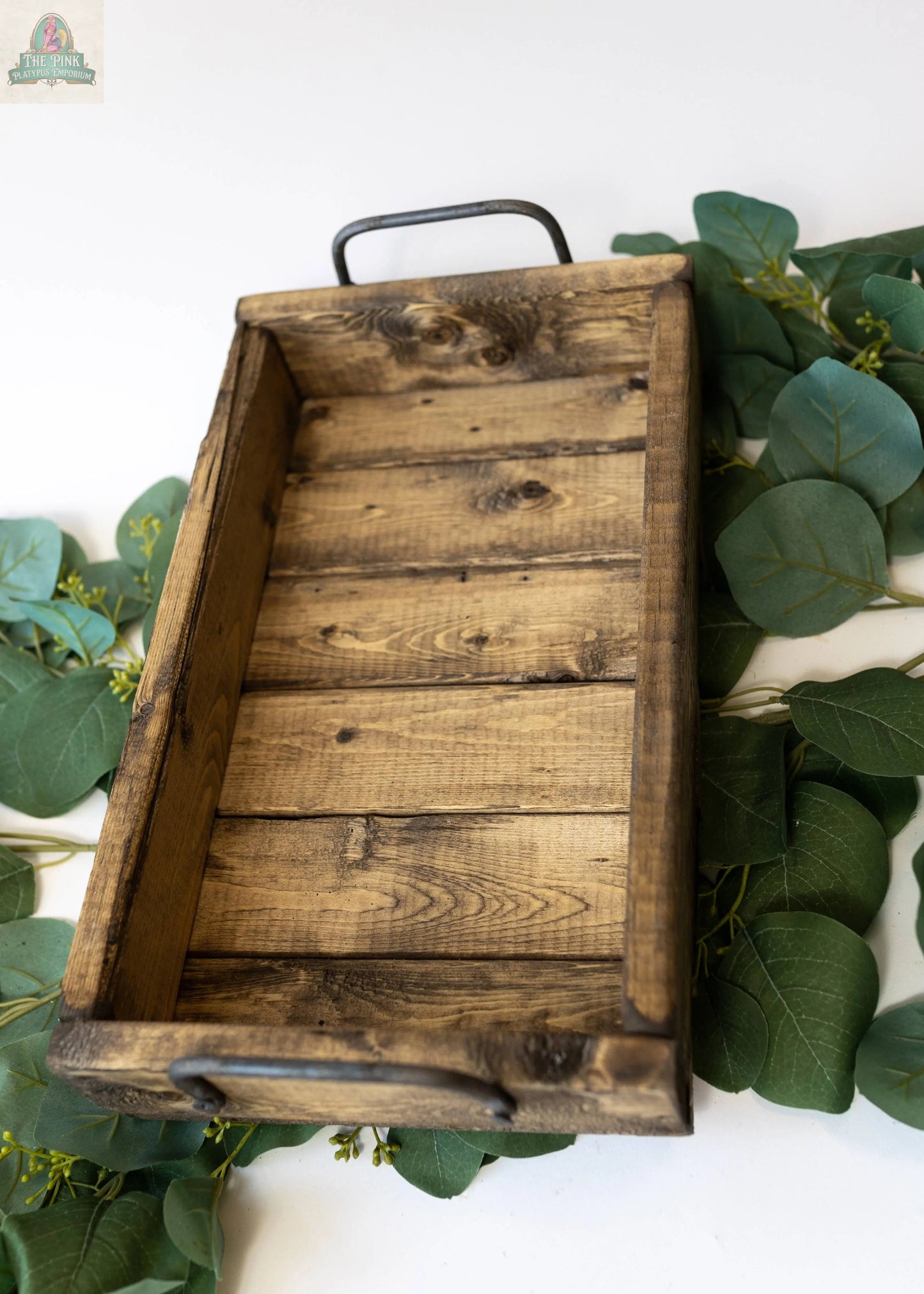 The Small Wood Tray: Distress Tray (14.5" x 8.5" x 3.25"), made from sustainable pine with metal handles, sits on a white surface and is surrounded by green eucalyptus leaves.