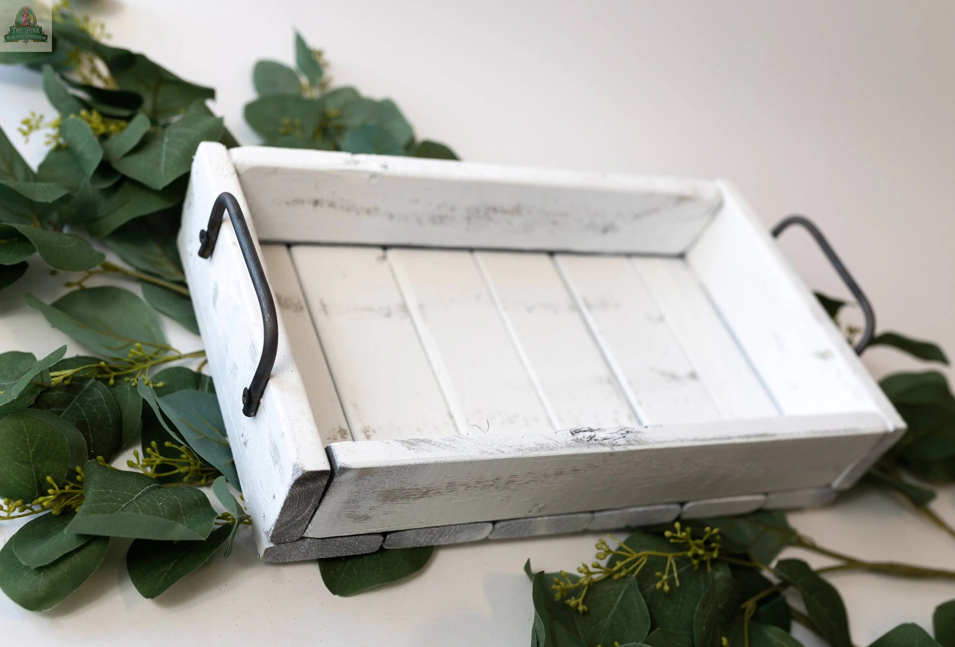 The Small Wood Tray: Distress Tray (14.5" x 8.5" x 3.25") is a rustic piece crafted from sustainable pine with black metal handles. Made in America, it features a vintage, weathered look and is perfect for displaying decor among greens.