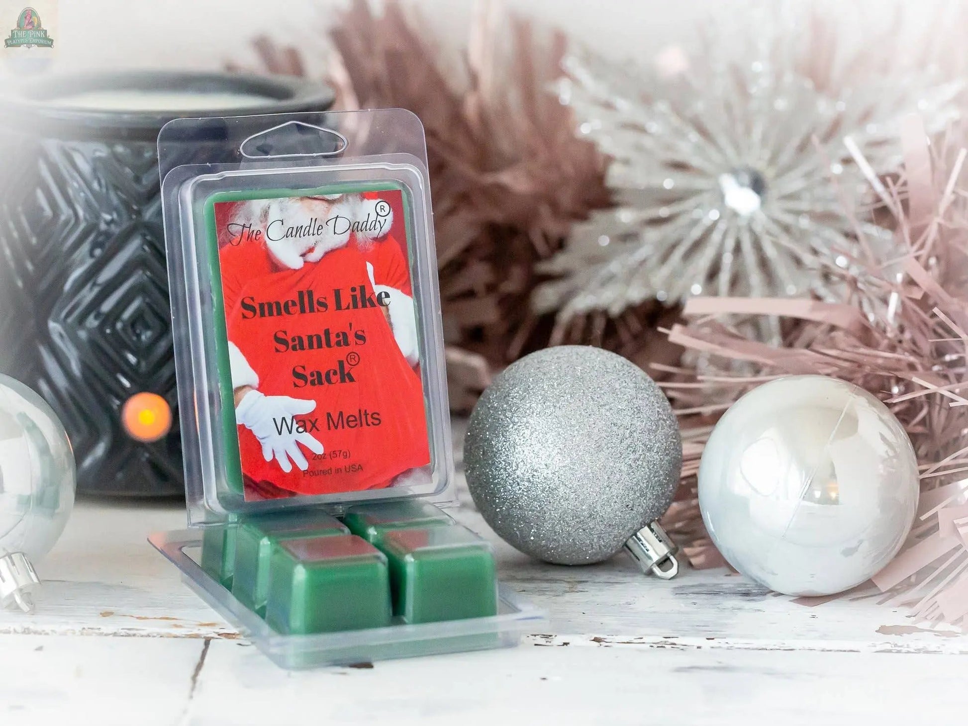A pack of "Smells Like Santa's Sack - Christmas Brown Sugar Fig" wax melts sits on a white surface next to silver and white ornaments, pink tinsel, and a black wax warmer, offering a cozy brown sugar fig scent.