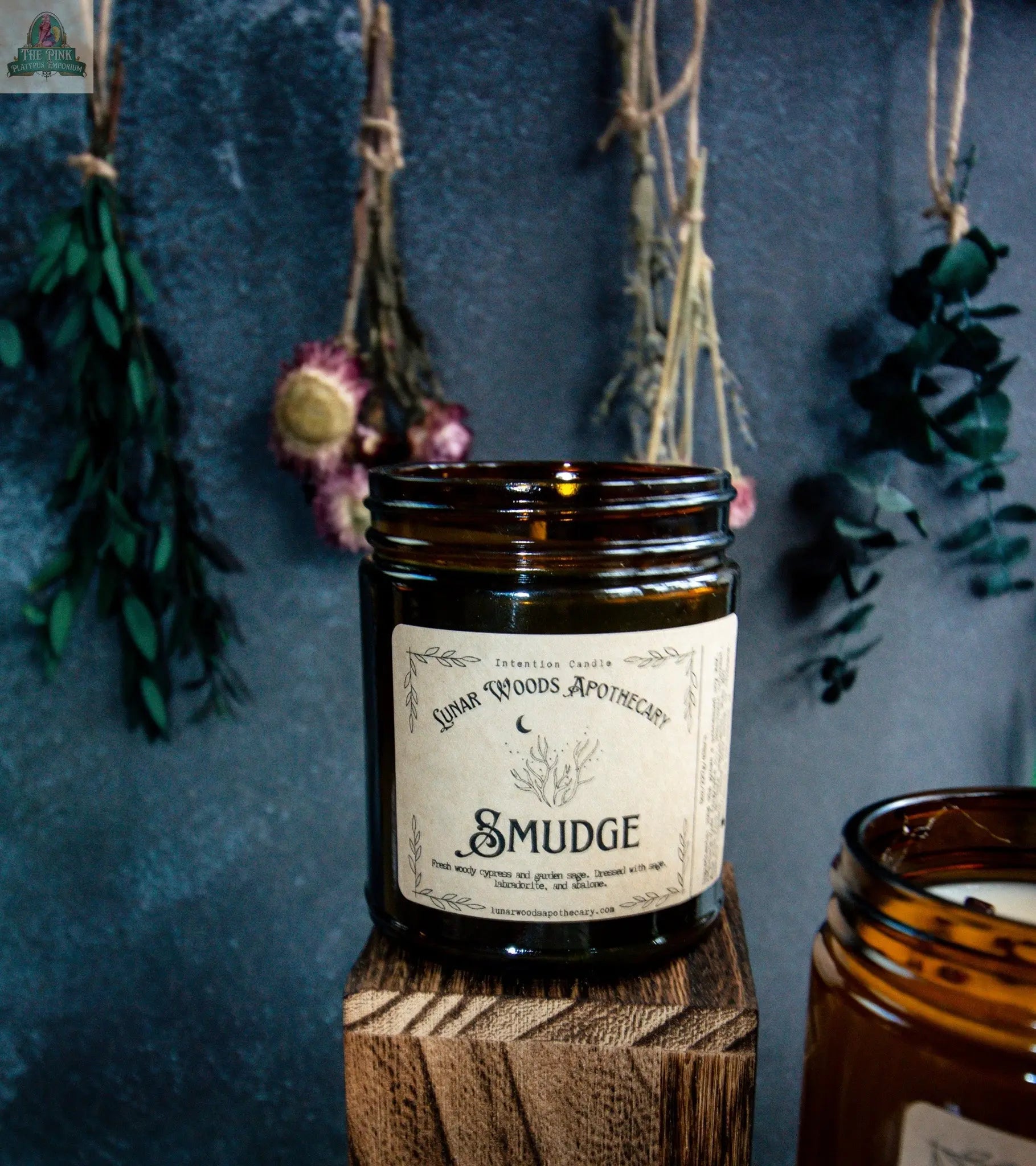 The Smudge amber glass candle sits on a wooden block. Behind it, dried flowers and herbs hang from strings against a dark wall. Another Smudge metaphysical candle is partly visible on the right.