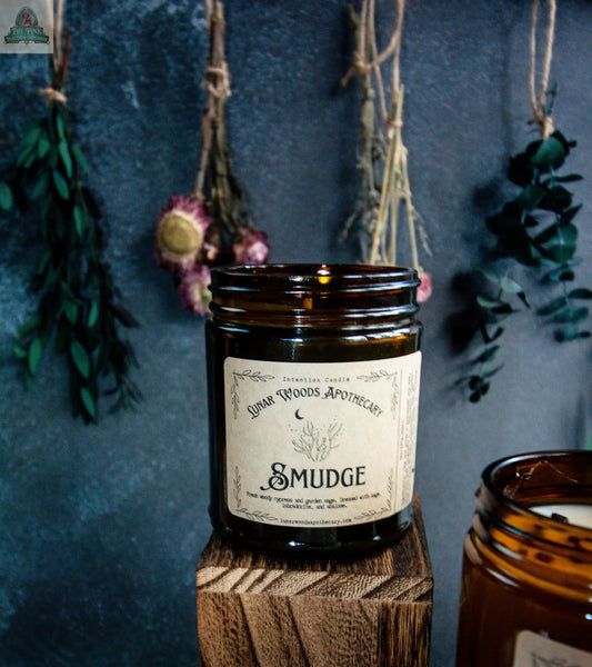 The Smudge amber glass candle sits on a wooden block. Behind it, dried flowers and herbs hang from strings against a dark wall. Another Smudge metaphysical candle is partly visible on the right.