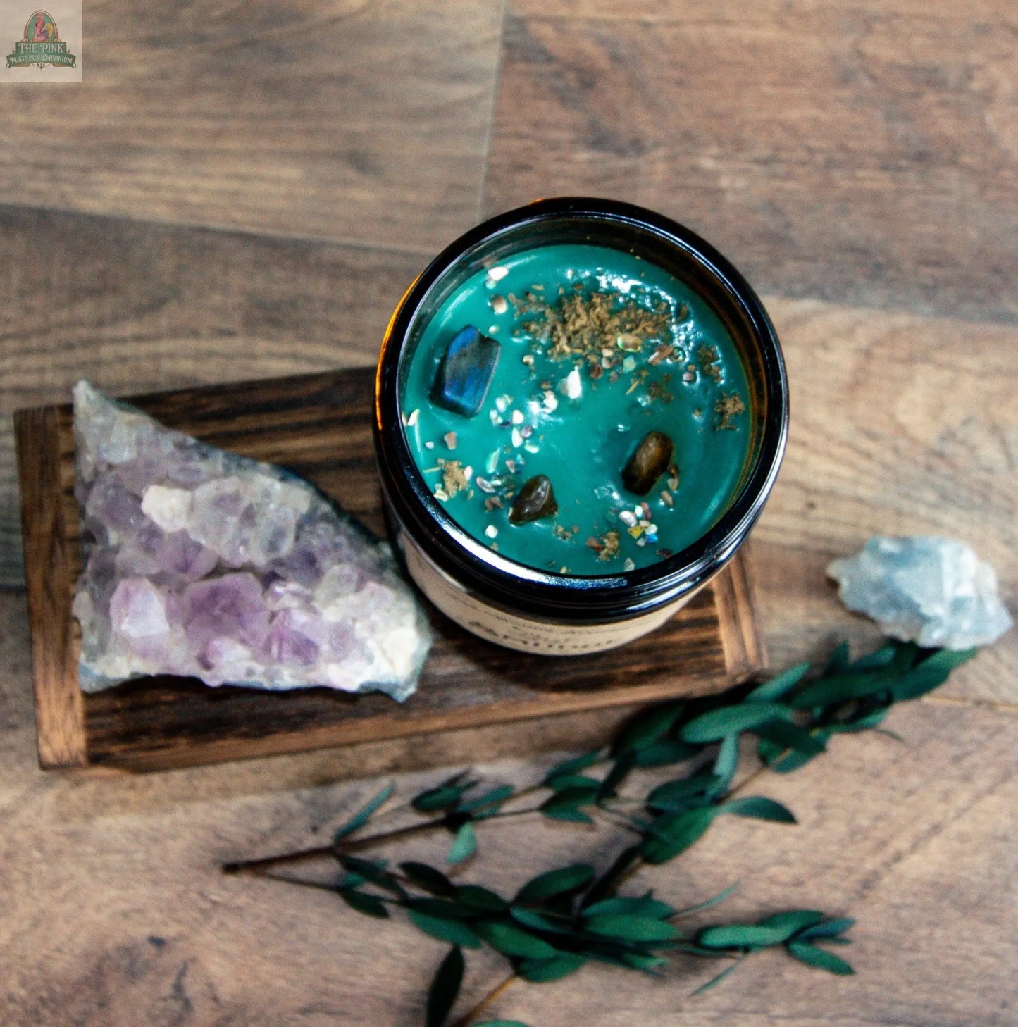 The Smudge candle, a blue-green metaphysical candle with herbs and small stones, rests on wood next to an amethyst cluster, a white stone, and greenery.