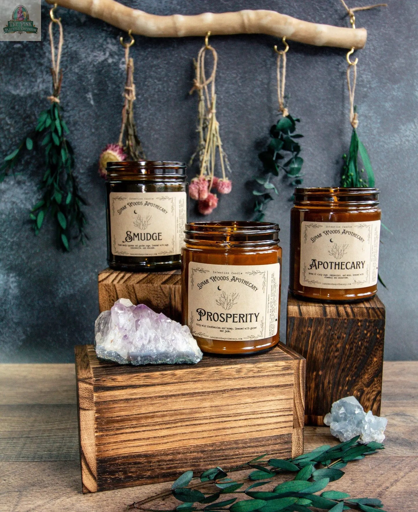 The amber Smudge candle sits on a wooden block surrounded by crystals and greenery, while dried herbs and flowers hang in the background, creating a mystical vibe for this metaphysical candle.