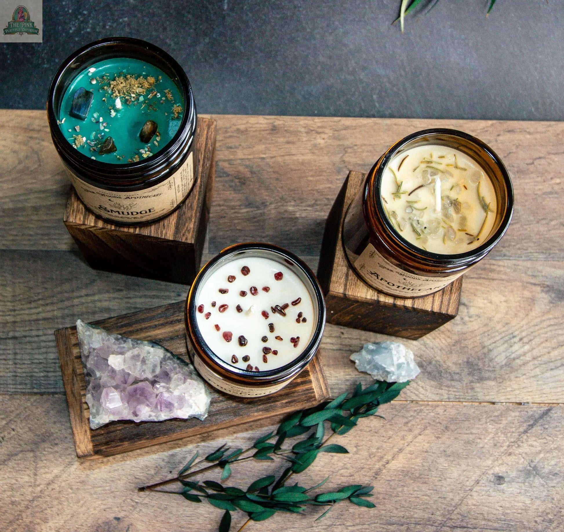 Three Smudge glass jar candles—including a sage candle topped with dried herbs, a green metaphysical candle with stones, and a white candle with peppercorns—sit on wood blocks, surrounded by crystals and greenery.