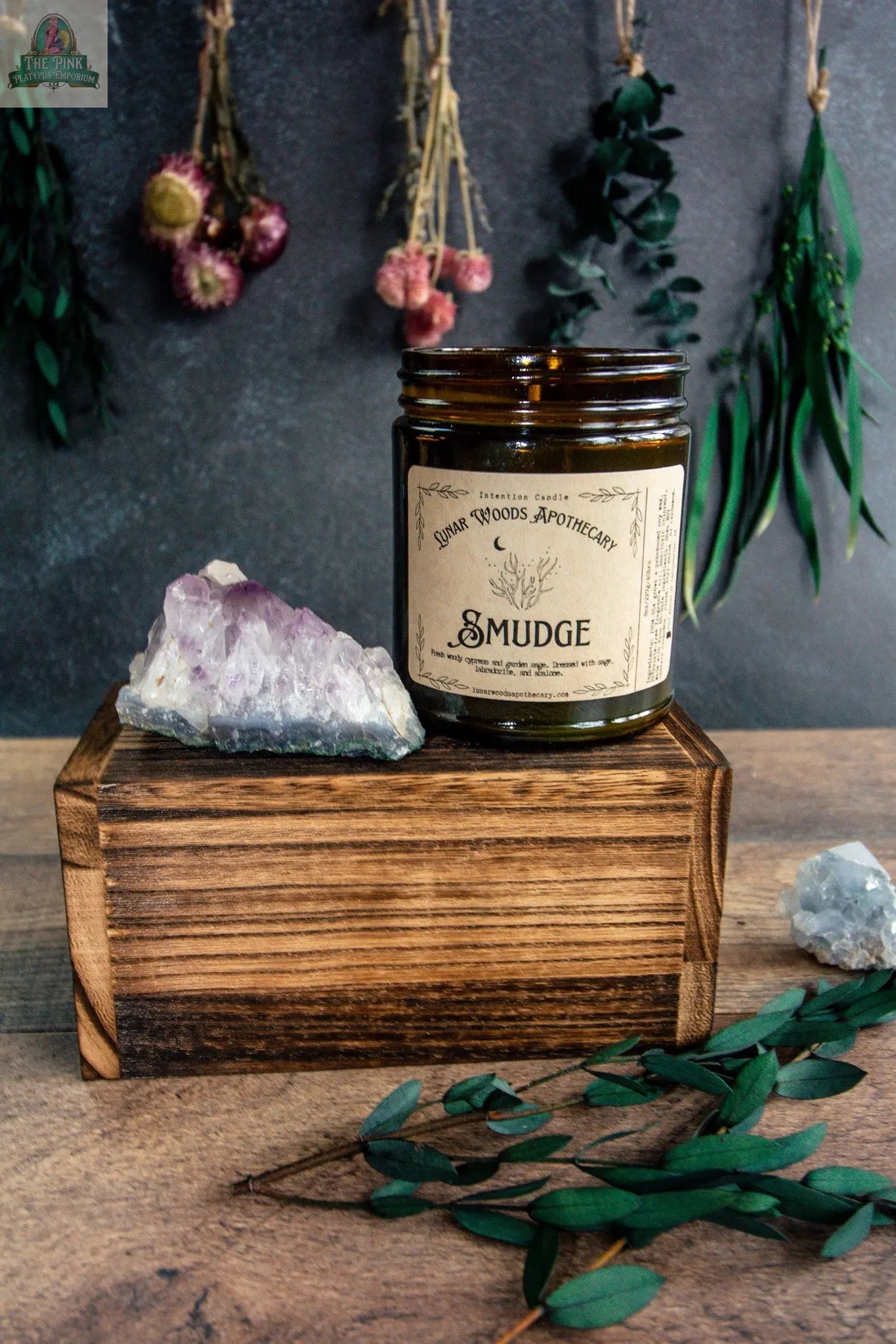 A brown glass jar labeled “Smudge” rests on a wooden box beside amethyst crystals, greenery, and dried flowers on a dark background—perfect for displaying your favorite Smudge candle or enhancing any candle ritual.