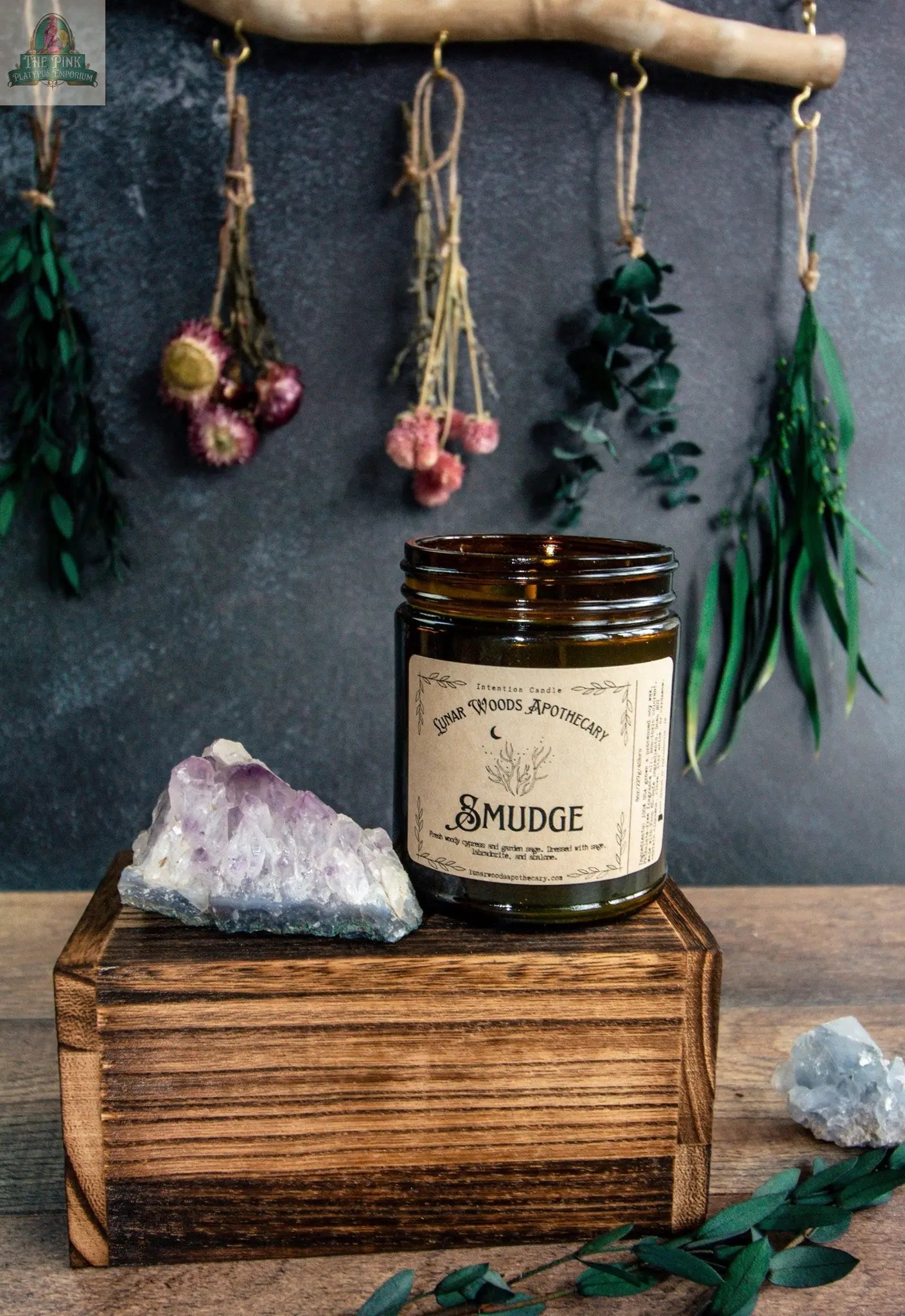 A brown glass jar labeled "Smudge" sits on a wooden box beside a purple crystal. Dried herbs and flowers hang from a branch in the background, while green leaves are scattered on the surface.