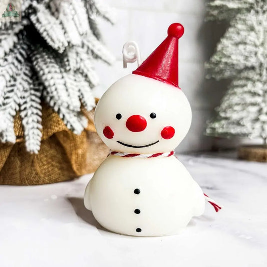 The Snowman Candle features a festive design with a red hat, rosy cheeks, nose, and a red-and-white scarf. Set on a snowy base among evergreens, it's ideal for winter decor or as a charming holiday gift.