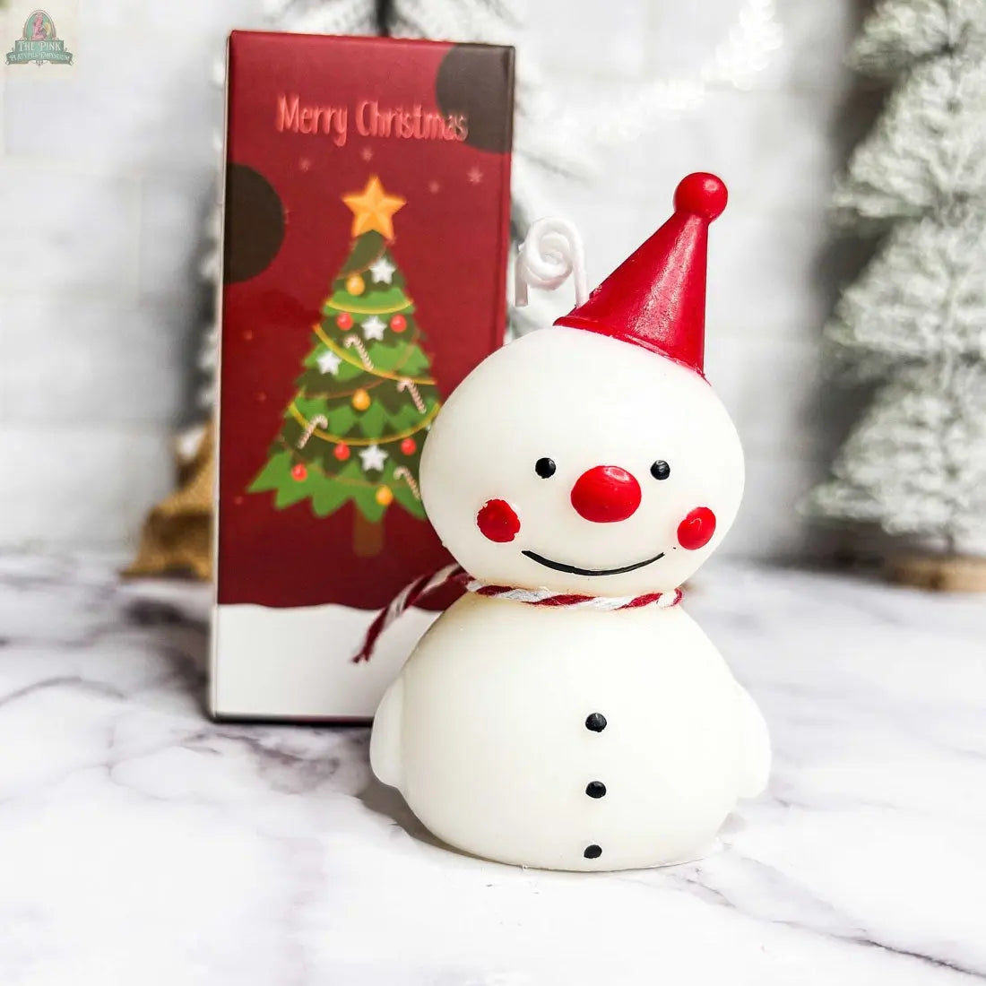 The Snowman Candle features a smiling snowman in a red party hat and scarf, standing before a gift box with a Christmas tree and "Merry Christmas" text—ideal as winter home decor or a festive holiday gift. Blurred decorations add a cozy touch.