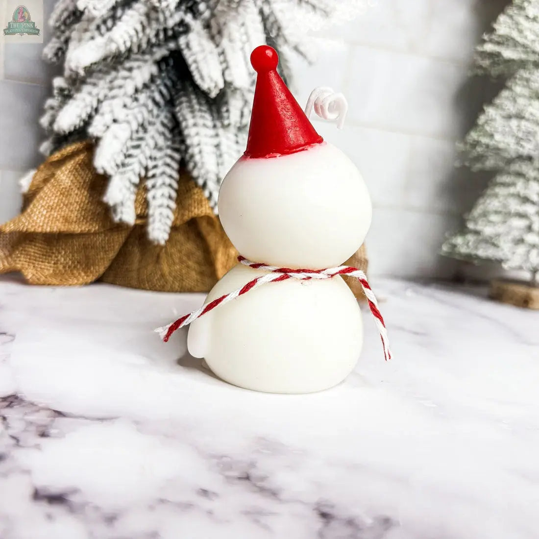 A Snowman Candle with a red party hat and a red-and-white string scarf sits on a marble surface, creating a charming holiday gift scene alongside snowy, burlap-wrapped trees in the background.